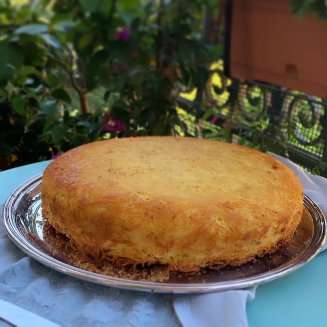 Torta dorata su un piatto argentato, circondata da piante verdi e fiori.